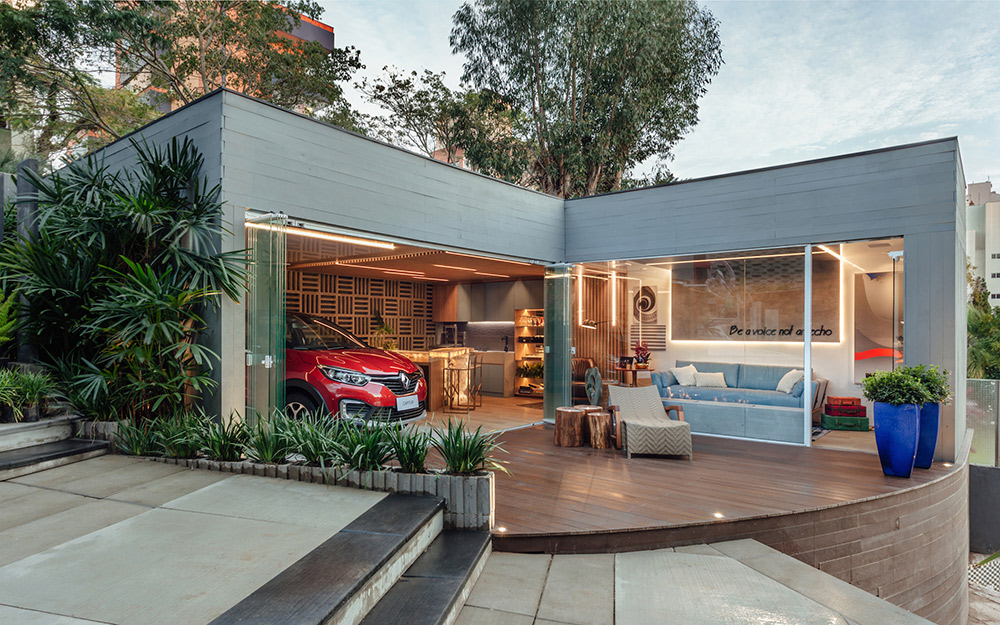 Garagem Renault - Cristina Da Luz e Davi Alexandre Heissler. O espaço é único e integrado, com fachada em pele de vidro que permite visualizar o lounge gourmet e o estar. Para surpreender, a dupla aposta no delicado azul do sofá de Pedro Mendes, em detalhes dourados e no mármore iluminado da bancada. A iluminação é destaque e se infiltra nos ripados de MDF, um dos vários materiais sustentáveis utilizados no projeto.