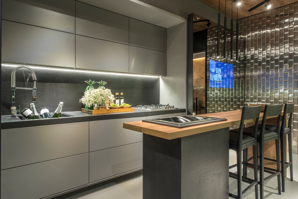 Cozinha Conceito - Ana Míriam Machado e Janice Diógenes. A dupla apostou no Dekton para modernizar o piso e as paredes. A esta base, agregaram o mobiliário cinza com portas de vidro prata. Além de automatizadas, as portas camuflam a geladeira, os fornos e as louças. A bancada recebe um tampo em Carvalho, mesma madeira que aquece o teto.