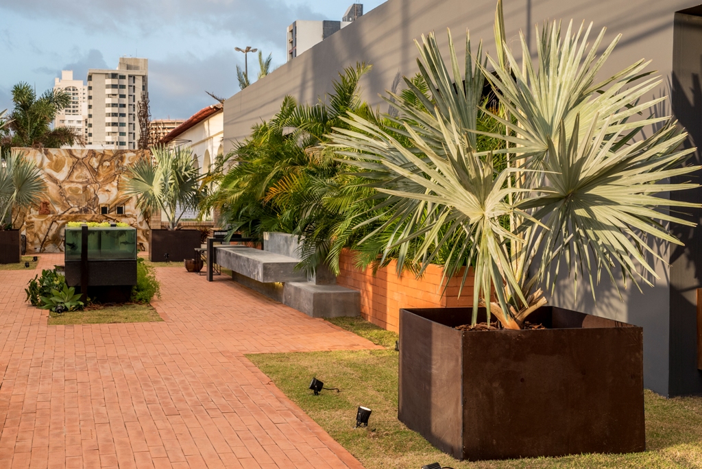 Elemental Garden - Michelle Viana. A imensa placa em mármore, com um desenho único, impacta ao primeiro olhar. O espaço também traz um quê de brutalismo, com o banco em pedra e as formas retilíneas. As plantas se moldam ao redor, nesta sala ao ar livre.