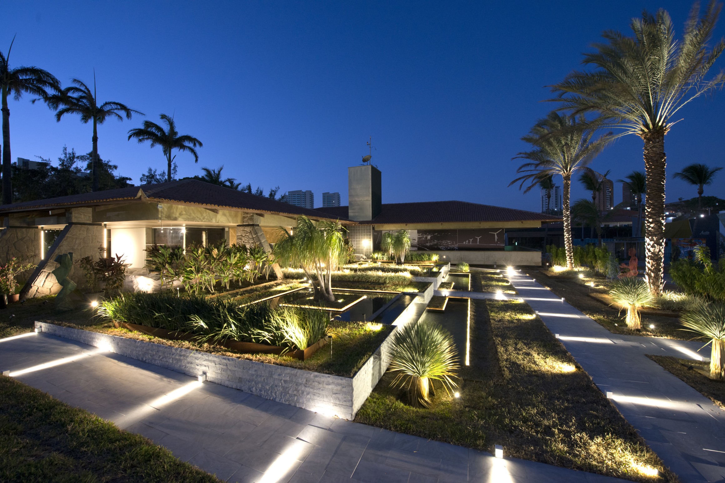 Jardim dos Ventos - Thiago Borges. Pedras naturais e granitos são os revestimentos, além de aço para as contenções, para um ar contemporâneo. Simétrico, o layout organiza espécies exóticas - algumas de grande porte, como palmeiras, tamareiras com 10m de altura e as patas-de-elefante com 25 anos de idade. A composição ainda reúne espelhos d’água de borda infinita, revestidos em pedra natural na cor preta.