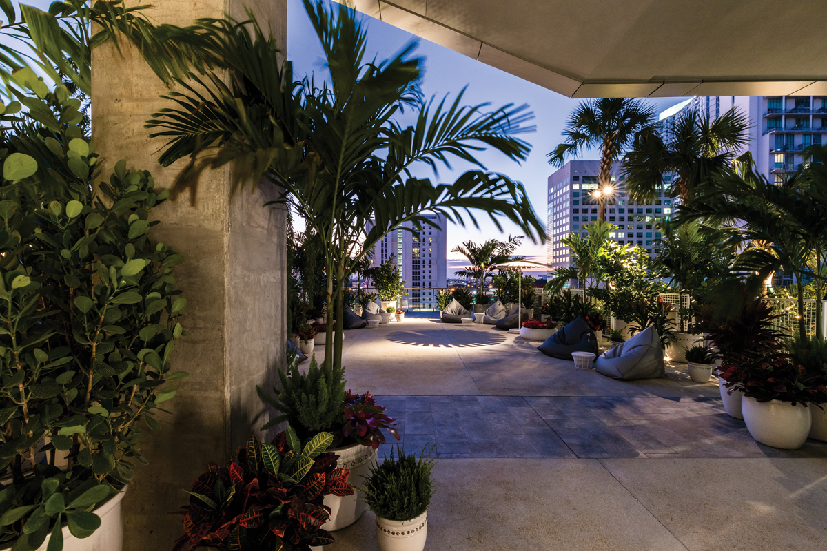 The Garden Bar - Adriana Mavignier Madeira. A vastidão do terraço inspirou a criação de áreas menores e protegidas, conectadas pela flora. Palmeiras, filodendros e samambaias foram mesclados, utilizando vasos de diversas alturas. Tudo para criar perspectivas confortáveis para os convidados que circulam ou aproveitam os pufes, enquanto conversam e contemplam o horizonte de Miami.