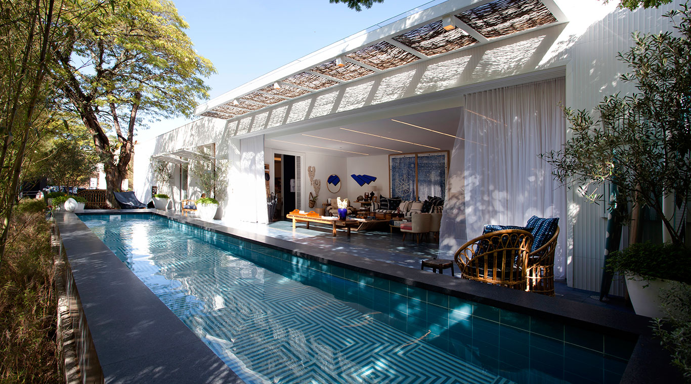 CASACOR São Paulo. Marina Linhares - Casa da Praia. Branco e matizes de azul trazem um quê de mar e de verão, em uma conversa perfeita com o tom natural da madeira e da palhinha. Móveis de junco, como os utilizados nas colônias inglesas no Caribe, foram desenhados pela Armando Cerello. Para completar a atmosfera litorânea, nada melhor do que uma piscina logo diante da sala.