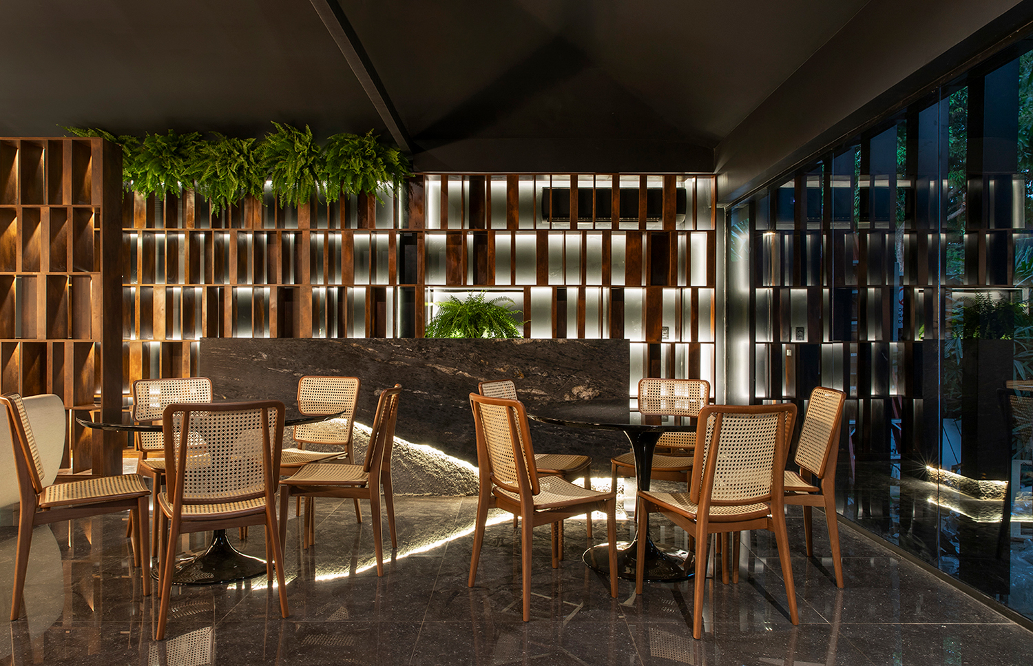 Restaurante - Poligonus Arquitetura. Concreto bruto, mármore e brises no tom de ouro velho revelam a vocação elegante e a personalidade do espaço, onde é possível conversar e degustar sob uma luz baixa, aconchegante. As cadeiras em madeira e palhinha reforçam o apelo natural. O vidro escuro traz um contraste com sua leveza e permite a visão da área externa, sem quebrar o clima intimista.