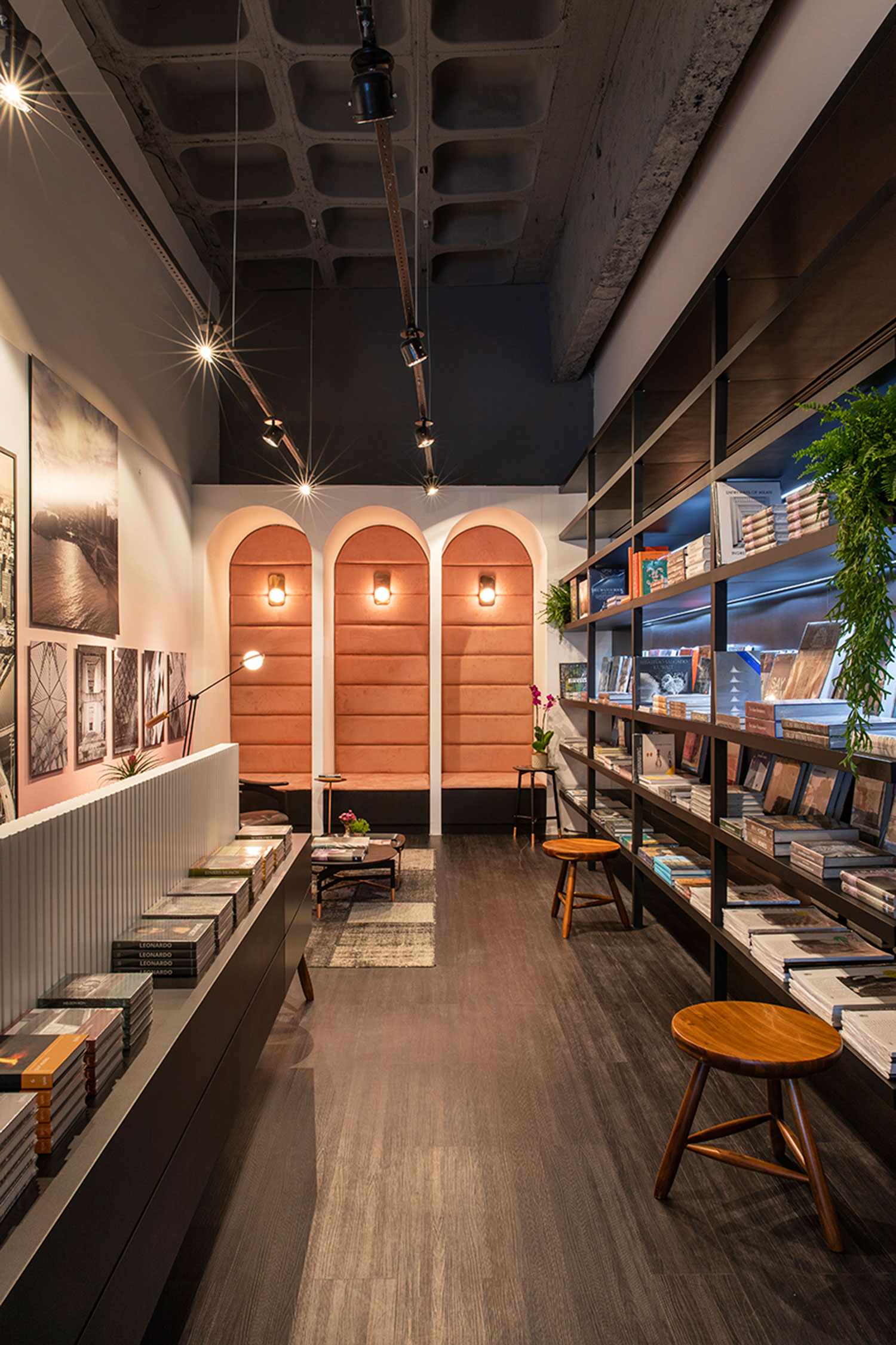 Art Bookshop - Leo Shehtman. O arquiteto e designer se baseia nas linguagens modernistas e pós-modernistas, como em Le Corbusier e Mondrian. Com 56 m², sua Livraria Viva resgata o recurso dos arcos nos vãos que formam o espaço de estar. De um lado, as fotografias de Marcelo Aniello são expostas na estrutura em serralheria que dita a organização. No oposto, a estante em ébano reúne o acervo de livros em sintonia com vegetações.