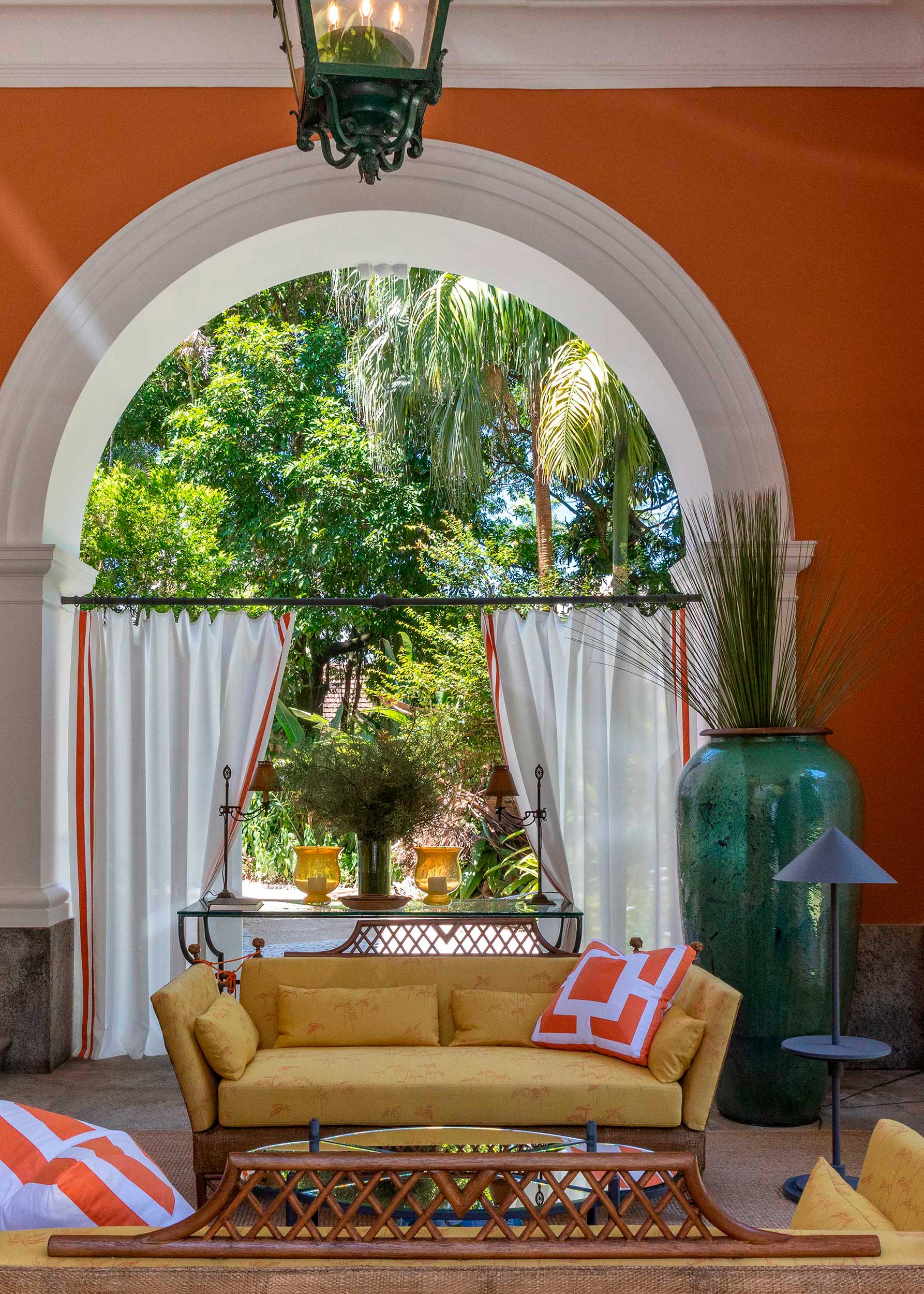 Atrium - CASACOR Rio de Janeiro 2021. Pensado como uma varanda intimista em que se pudesse aproveitar a bela vista para os amplos jardins da residência, o antigo porte-cochère da casa ganhou ambientação casual chic com toques tropicais e o uso de materiais naturais como fibras e palha. Arcos brancos enquadram a natureza em volta.