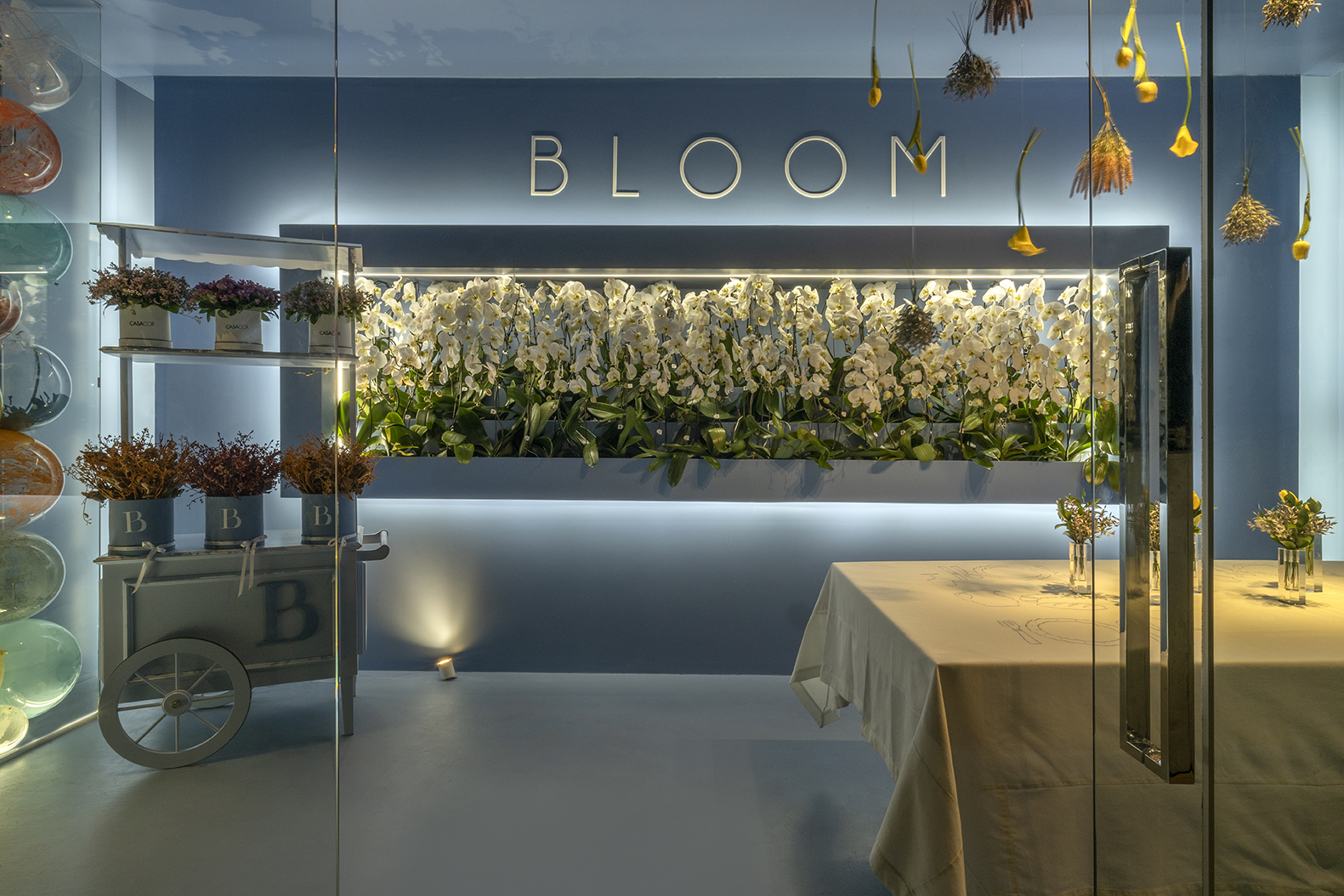 Roberto Guedes - Bloom. Um dos ambientes mais instagramáveis da mostra, Roberto Guedes projetou uma parede de balões e flores excêntricas. Iluminação de led dá o charme ao ambiente minimalista. A inspiração do arquiteto foi em lojas italianas ligadas à área de moda e flores, após uma longa pesquisa na forma como estabelecimentos desses nichos apresentam suas criações. Com 18 m² em construção modular, o ambiente utiliza a cor azul tibet. Usando a mesma cor em todo o ambiente com o intuito de levar o visitante a valorizar apenas um momento, o espaço traz ornamentação com pratos exclusivos, vasos e flores.