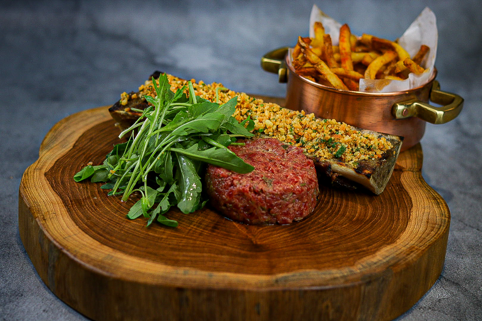 Steak tartar & tutano, uma barca de tutano grelhada com farofa de pão de queijo, steak tartar, batata frita e salada de rúcula.